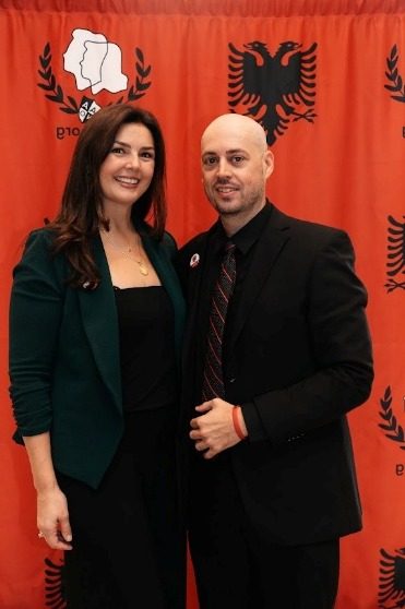 Two attendees in front of the red AANO backdrop with the Albanian double-headed eagle.