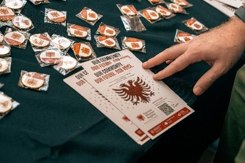 A hand reaching across a table of National Albanian Registry pins and a Count Me Albanian card.