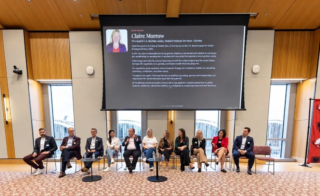 The full panel seated on stage as a panelist's biography shows on the screen.