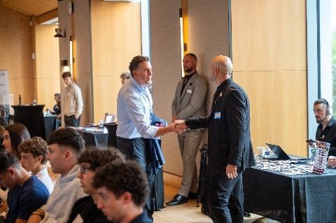 Two attendees shaking hands at the back of the room while students look on.